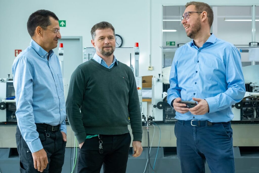 Khalid Dawam, Jan Schölzel und Ronny Richter stehen in einer modernen Werkstatthalle von PostPrint Leipzig und unterhalten sich. Sie tragen Hemden oder Pullover und wirken entspannt und freundlich. Im Hintergrund sind Maschinen und Arbeitsmaterialien zu sehen.